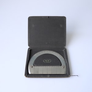 May include: A vintage silver-toned protractor with degree markings, housed in an open, dark gray case. The protractor has a semi-circular shape with a straight edge and is marked with numbers and lines. The case is lined with a soft, dark gray material.