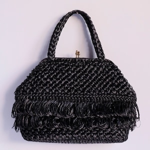 May include: A black woven handbag with a braided handle and fringe detail. The bag features a clasp closure and a structured shape. The handbag is likely made of a natural fibre material, suitable for carrying essentials.