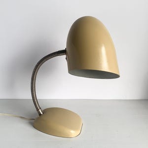 Puede incluir: Lámpara de escritorio vintage con pantalla y base redondeadas de color crema. El cuello de cisne flexible plateado permite una iluminación ajustable. La lámpara está diseñada con una estética minimalista.