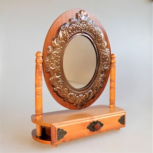 May include: A wooden vanity mirror with an oval-shaped mirror and ornate bronze-colored frame. The mirror is set on a wooden base with a drawer and decorative hardware. The mirror is approximately 18 inches tall.