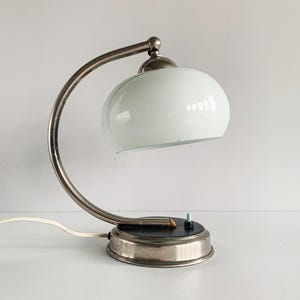 May include: A vintage-style desk lamp with a curved, silver-toned metal arm and a round, white glass shade. The base is a dark color with a silver rim and a small switch. The lamp is plugged in with a white cord.
