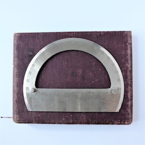 May include: A vintage metal protractor with degree markings, resting on a rectangular, dark burgundy book. The protractor is a semi-circle shape with a ruler along the straight edge. The book has a worn, textured appearance.