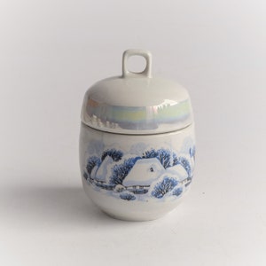 May include: A ceramic jar with a lid, featuring a winter scene with snow-covered houses and trees in blue and white. The lid has a small loop handle. The jar has a pearlescent band around the middle.