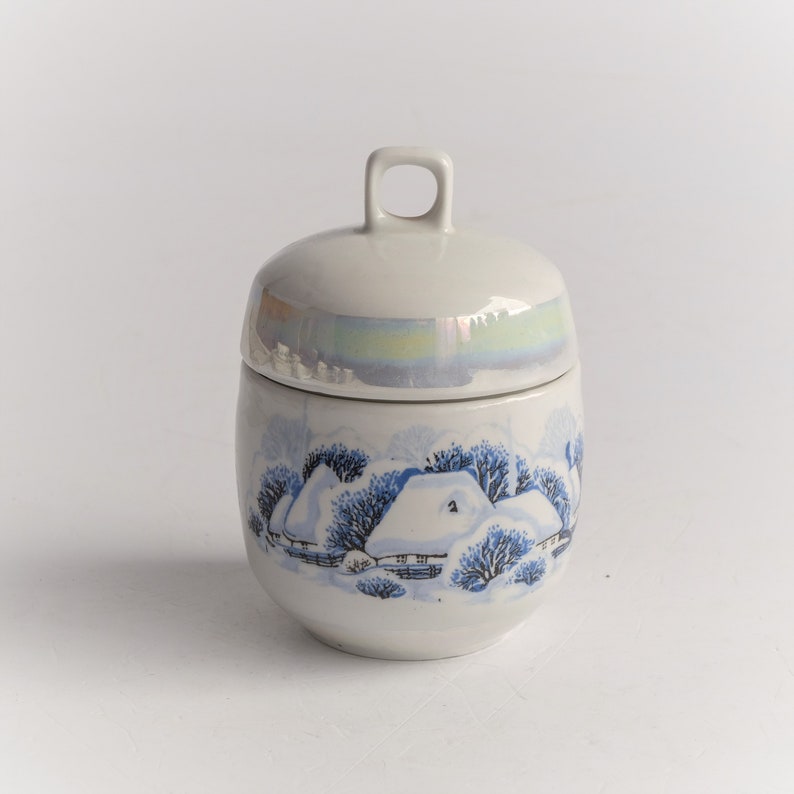 May include: A ceramic jar with a lid, featuring a winter scene with snow-covered houses and trees in blue and white. The lid has a small loop handle. The jar has a pearlescent band around the middle.
