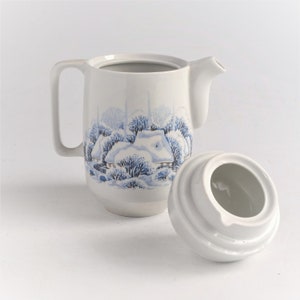 May include: White ceramic creamer with a winter scene design in blue. The creamer has a handle and a spout, and the lid is off to the side. The winter scene depicts a snow-covered cottage and trees.