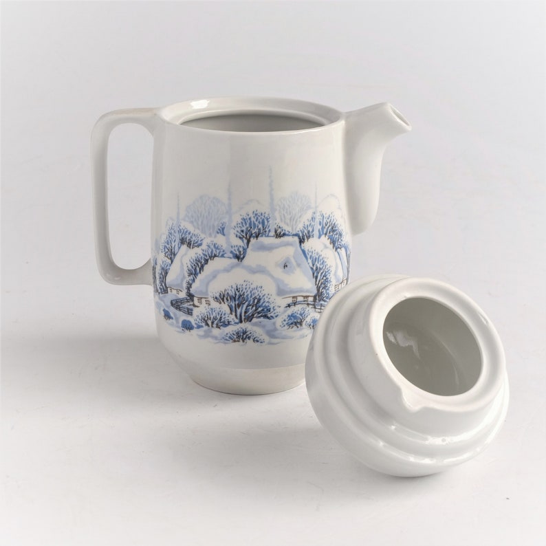 May include: White ceramic creamer with a winter scene design in blue. The creamer has a handle and a spout, and the lid is off to the side. The winter scene depicts a snow-covered cottage and trees.