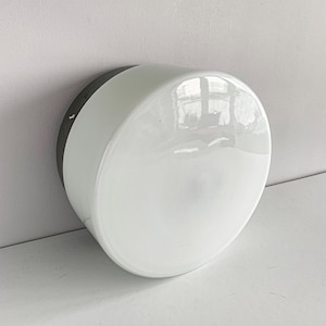 Könnte beinhalten: Eine weiße, halbkugelförmige Wandleuchte mit dunkelgrauem Rand. Die Leuchte hat eine glänzende Oberfläche und ist an einer weißen Wand montiert. Die Leuchte ist ein modernes Design.