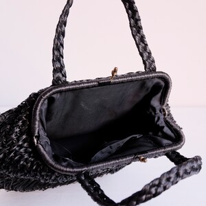 May include: Black woven handbag with plaited handles and a clasp closure. The interior is lined with black fabric and has a small pocket. The bag has a vintage aesthetic.