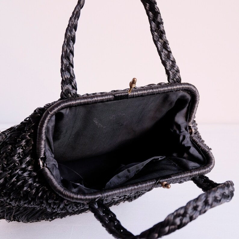 May include: Black woven handbag with plaited handles and a clasp closure. The interior is lined with black fabric and has a small pocket. The bag has a vintage aesthetic.