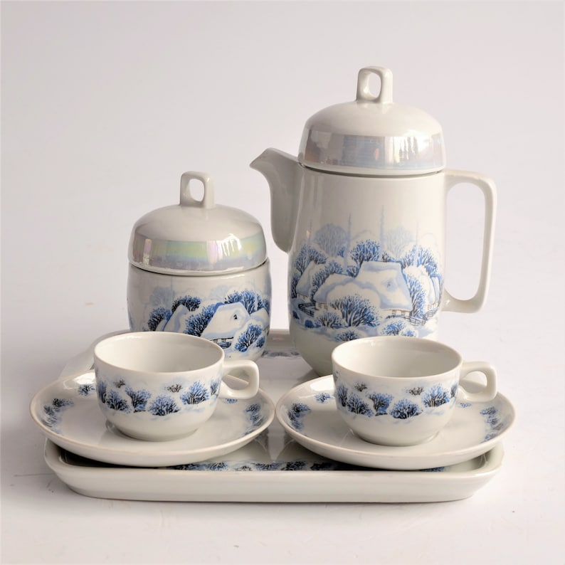 May include: A white ceramic tea set with a winter landscape design. The set includes a teapot, sugar bowl, two cups and saucers, and a rectangular tray. The design features blue trees and snow-covered houses. The set is on a white background.