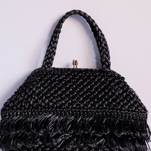 May include: Black woven handbag with a braided handle and fringe detail. The bag has a brass clasp and a textured, chevron-like weave. The design is vintage-inspired, suitable for various occasions.