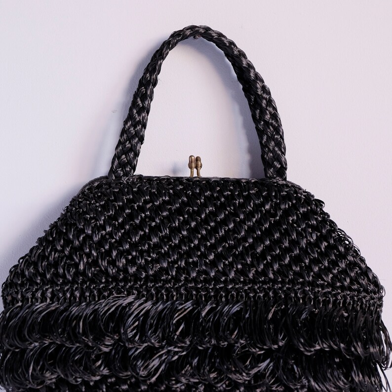 May include: Black woven handbag with a braided handle and fringe detail. The bag has a brass clasp and a textured, chevron-like weave. The design is vintage-inspired, suitable for various occasions.