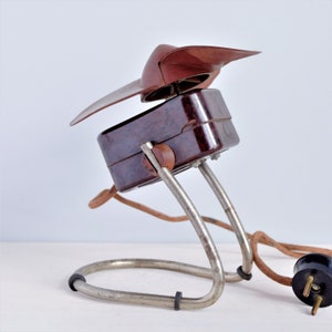 May include: A vintage brown electric fan with a unique blade design. The fan has a rectangular body, supported by a curved metal stand. The power cord is brown with a black plug. The fan's blades are a dark brown color.