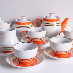 Könnte beinhalten: Ein weißes Keramik-Teeservice mit orangefarbenen und goldenen Akzenten. Das Set beinhaltet eine Teekanne, eine Zuckerdose, einen Sahnekännchen, Tassen und Untertassen. Die Tassen und Untertassen haben orangefarbene und goldene Streifen.