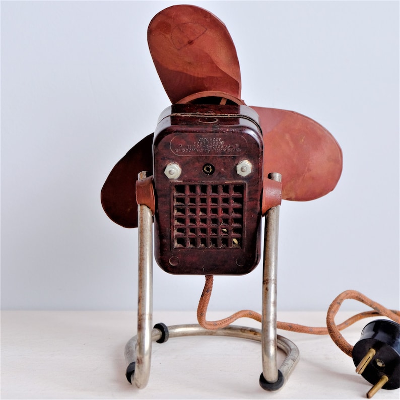 May include: Vintage brown electric fan with two blades. The fan has a dark brown body, a metal stand, and a brown power cord with a black plug. The fan's blades are a lighter shade of brown.