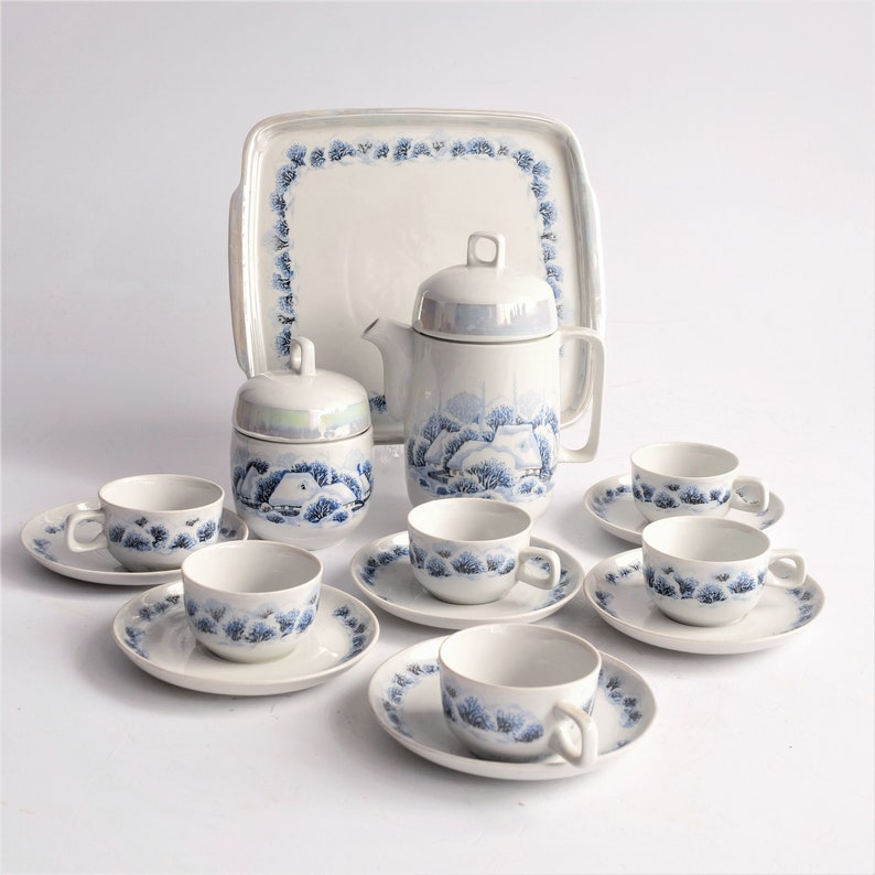 May include: A vintage white porcelain tea set with a winter landscape design. The set includes a teapot, sugar bowl, serving tray, and six teacups with saucers. The design features blue trees and houses.