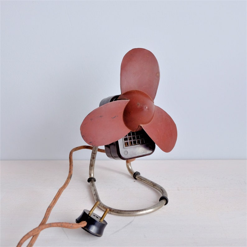 May include: An antique electric fan with a maroon propeller and a dark brown motor housing. The fan has a metal frame and a brown, cloth-covered power cord with a black plug. The fan is sitting on a white surface.