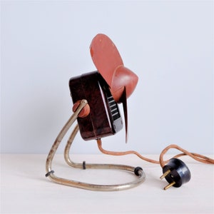 May include: Vintage brown and red electric fan with a metal stand. The fan has a dark brown body, a red fan blade, and a metal stand. The power cord has a black plug.