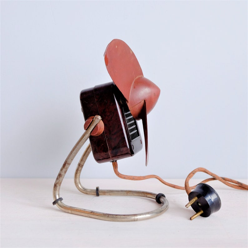 May include: Vintage brown and red electric fan with a metal stand. The fan has a dark brown body, a red fan blade, and a metal stand. The power cord has a black plug.