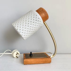 May include: A vintage desk lamp with a white, diamond-patterned lampshade and a wooden base. The lamp has a curved brass-coloured arm and a wooden top. It features a black on/off switch and a white electrical plug.
