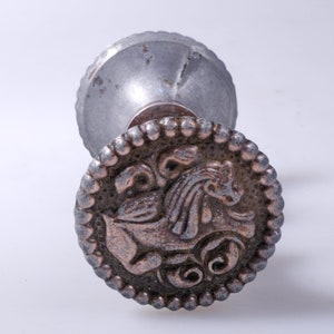 May include: A vintage silver-toned door knob with a detailed design. The knob features a circular face with a raised relief of a mythical creature, surrounded by a beaded border. The top portion is a fluted, rounded shape.