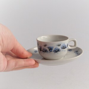 May include: A white ceramic teacup and saucer set. The cup and saucer are decorated with a blue and black tree design. The cup has a small handle. The set is held by a hand against a white background.