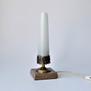 May include: A vintage table lamp with a white, cylindrical shade. The lamp features a brass base with decorative detailing and a square, brown stone base. The lamp is plugged into a white wall outlet.