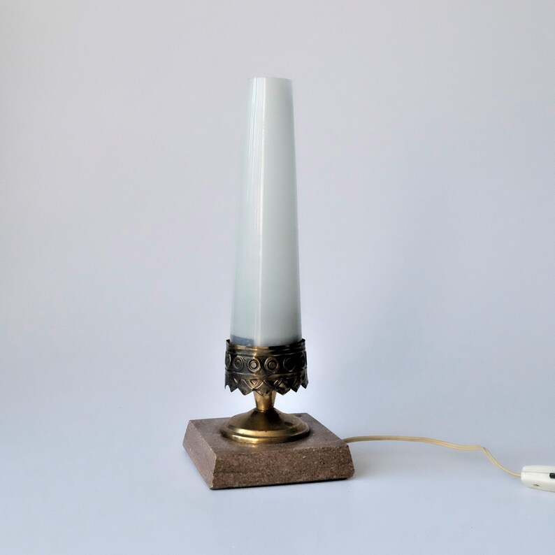 May include: A vintage table lamp with a white, cylindrical shade. The lamp features a brass base with decorative detailing and a square, brown stone base. The lamp is plugged into a white wall outlet.