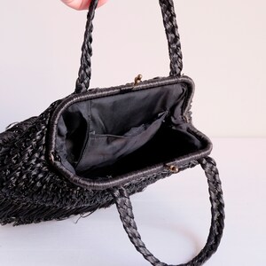 May include: Black woven handbag with plaited handles and a clasp closure. The interior is lined with black fabric and features a small pocket. The bag has a rectangular shape and a fringe detail along the bottom edge.