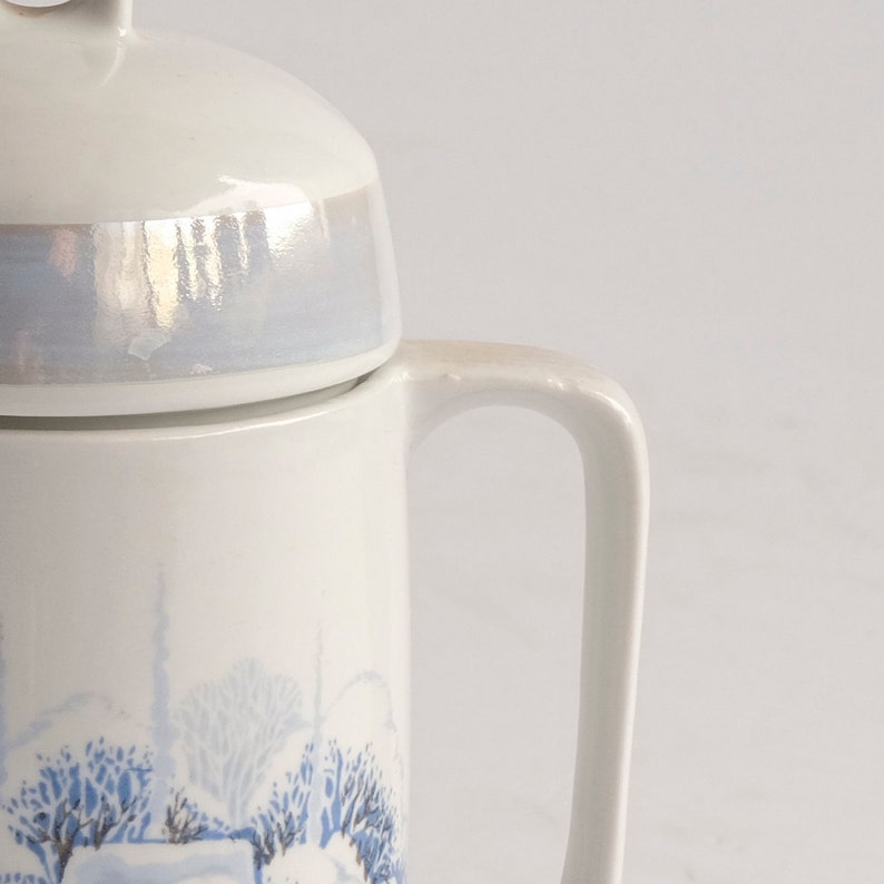 May include: White ceramic teapot with a lid and handle. The lid has a pearlescent finish. The body of the teapot features a blue and white winter landscape design. The handle is curved and attached to the side of the teapot.