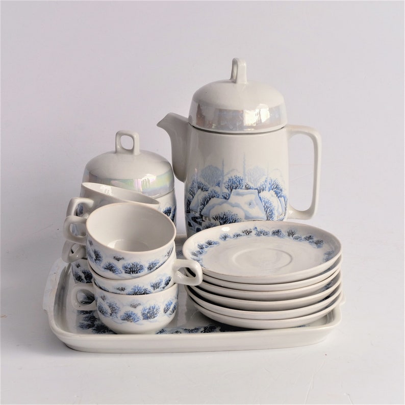 May include: A white ceramic tea set with a blue winter landscape design. The set includes a teapot, sugar bowl, cups, saucers, and a serving tray. The cups and saucers have a blue tree pattern around the edges.