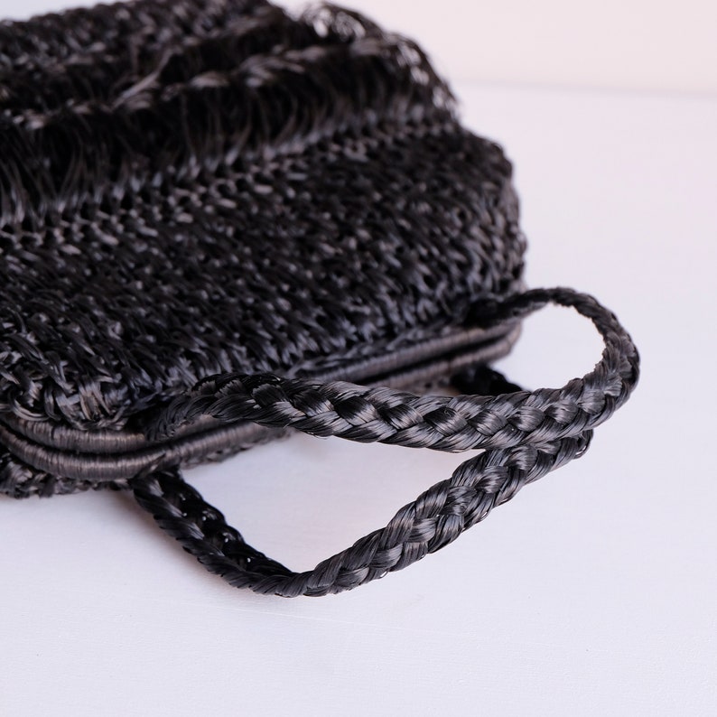 May include: A black woven handbag with a braided handle. The bag has a rectangular shape and a fringe detail at the top. The handle is thick and sturdy, and the bag appears to be made of a durable material.