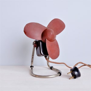 May include: Vintage electric fan with three maroon blades and a black motor housing. The fan has a chrome-colored metal stand and a brown power cord with a black plug. A classic home appliance.