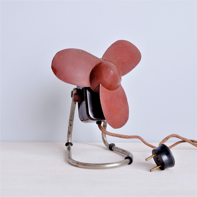 May include: Vintage electric fan with three maroon blades and a black motor housing. The fan has a chrome-colored metal stand and a brown power cord with a black plug. A classic home appliance.