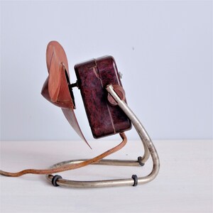 May include: An antique electric fan with a maroon-colored fan blade and a dark brown motor housing. The fan is mounted on a silver-colored, curved metal stand with a braided power cord. The fan is a vintage item.