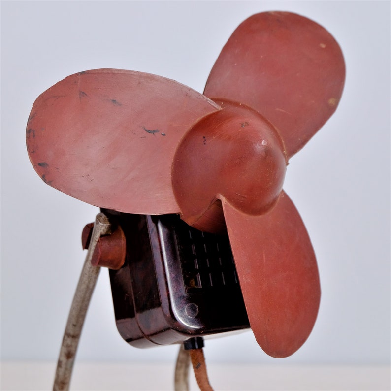 May include: A vintage electric fan with three reddish-brown blades. The fan's motor housing is dark brown, and the stand is made of metal. The fan is a classic design, likely from the mid-20th century.