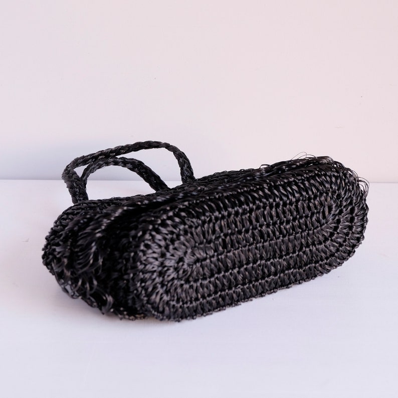May include: A black woven handbag with braided handles. The bag has a rectangular shape with rounded edges. The material appears to be a tightly woven synthetic fibre, giving it a textured appearance. The bag is set against a white background.