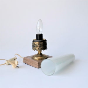 May include: Vintage table lamp with a brass base, decorative detailing, and a clear lightbulb. The lamp sits on a square, speckled base and includes a white, cylindrical lampshade. A power cord with a plug is also visible.