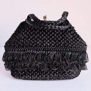 May include: Black woven handbag with a braided handle and fringe detail. The bag has a clasp closure and a structured shape. The fringe adds a decorative element to the bottom of the bag. The bag is made of a woven material.