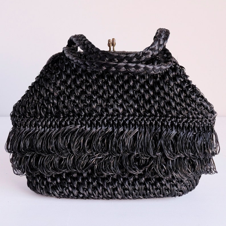 May include: Black woven handbag with a braided handle and fringe detail. The bag has a clasp closure and a structured shape. The fringe adds a decorative element to the bottom of the bag. The bag is made of a woven material.