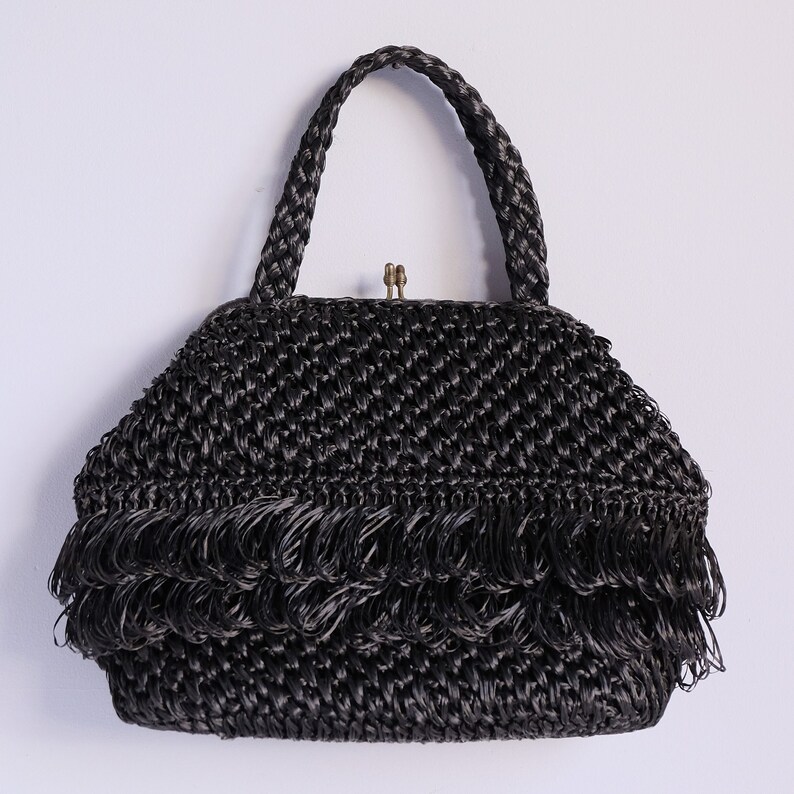 May include: A black woven handbag with a braided handle and a decorative fringe along the bottom. The bag has a clasp closure and a structured shape, suitable for carrying essentials.