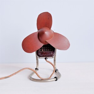May include: A vintage electric fan with a maroon propeller and housing. The fan has three blades and a metal frame with a brown power cord. The fan is designed to sit on a surface and has a retro aesthetic.