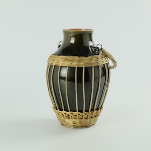 Pode incluir: Um vaso de cerâmica verde escuro com uma base de vime tecido e uma alça tecida. O vaso tem um acabamento brilhante e uma forma cilíndrica, com linhas verticais de vime ao redor do corpo. A alça é fixada com anéis de metal.