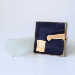 May include: A vintage lamp base with a rectangular, dark blue velvet-covered base and a cylindrical frosted glass shade. The base has a wooden keyhole detail and a small label. A power cord extends from the base.