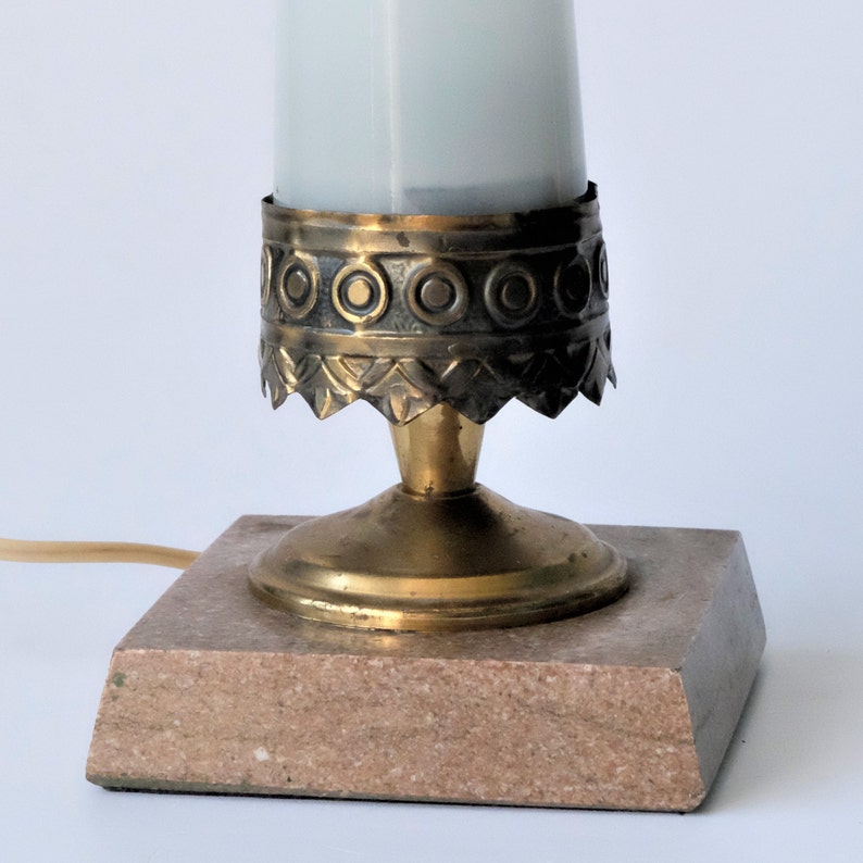 May include: A small, vintage table lamp with a frosted glass shade. The lamp features a brass base with decorative detailing and a square, pinkish-beige marble base. A power cord extends from the base.