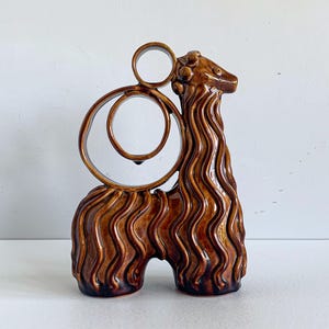 Op de afbeelding: Een bruin geglazuurd keramisch beeld van een gestileerd dier met een lange nek en een golvend, getextureerd lichaam. Het figuur heeft twee prominente cirkelvormige ringen die uit de rug steken, wat zorgt voor een open, geometrisch ontwerp.