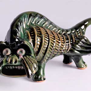 May include: A ceramic sculpture of a stylized creature in shades of green and brown. The sculpture has a large open mouth, prominent eyes, and a handle-like feature at the end. The piece is glazed and has a glossy finish.