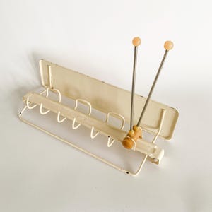May include: Vintage off-white metal wall-mounted coat rack with six hooks and a unique antenna-like design. Features two slender metal rods topped with peach-colored spheres. A charming retro piece for entryway organization.