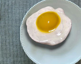 Può includere: Un piatto bianco con un uovo fritto sopra. Il tuorlo dell'uovo è giallo brillante e l'albume è cotto a una consistenza morbida. Il piatto è su un sottopiatto a strisce grigie e bianche.