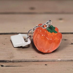 May include: A pumpkin-shaped keychain with a silver chain and a white accessory. The pumpkin is orange with a green stem. The keychain is on a wooden surface.